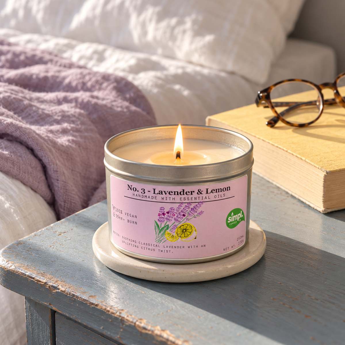 Lit Simpl Lavender & Lemon tin candle on a blue bedside table beside a book and glasses, with lavender bedding behind.