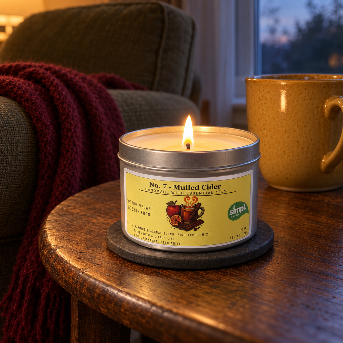 Lit Simpl Mulled Cider tin candle on a wooden side table beside a yellow mug, with a burgundy blanket and dusk outside.