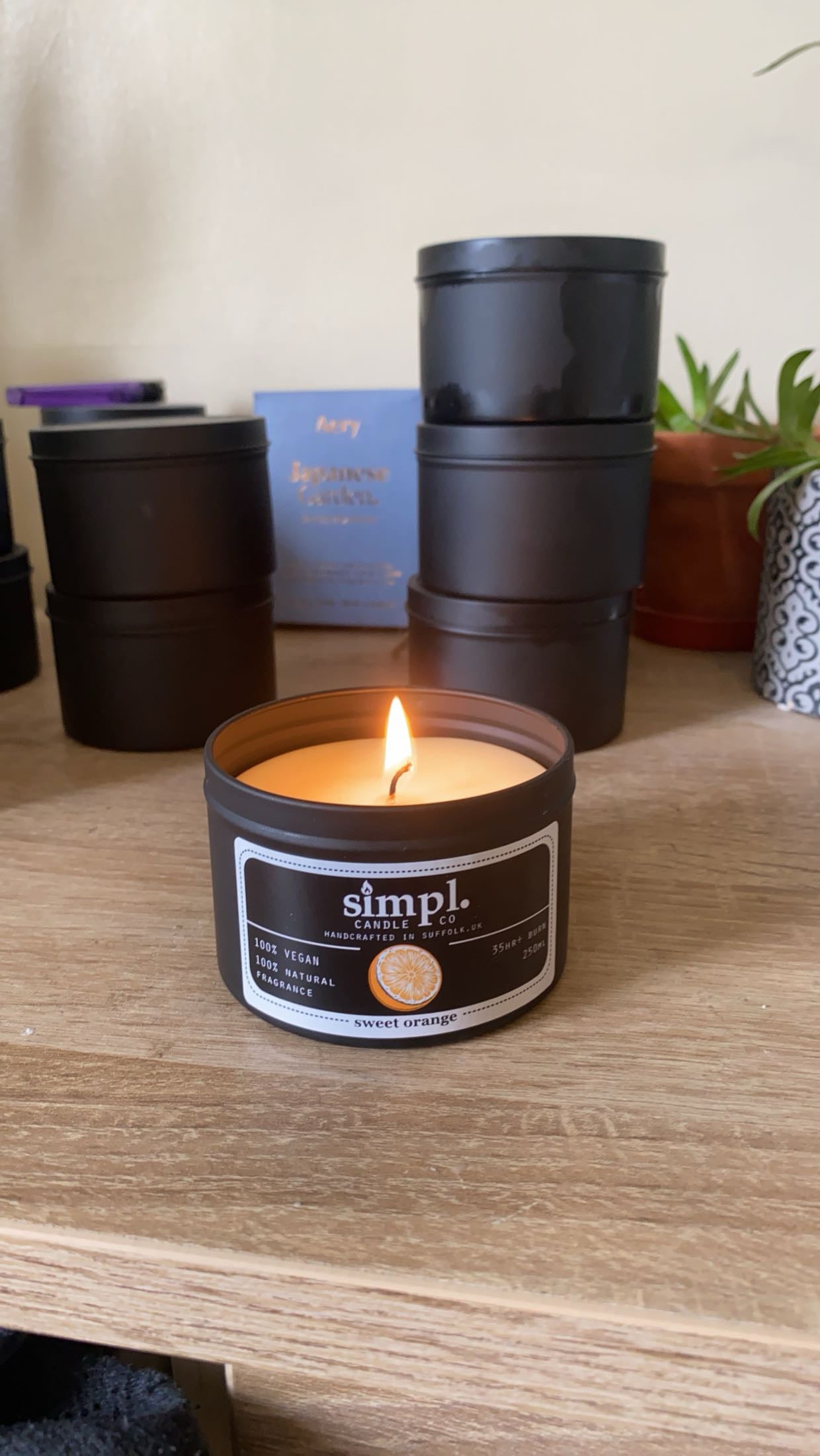 Candle in a 'simpl' tin on a wooden surface with other containers and a plant in the background.